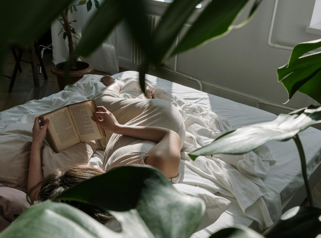 A woman lies in bed reading a book, surrounded by greenery and natural light.