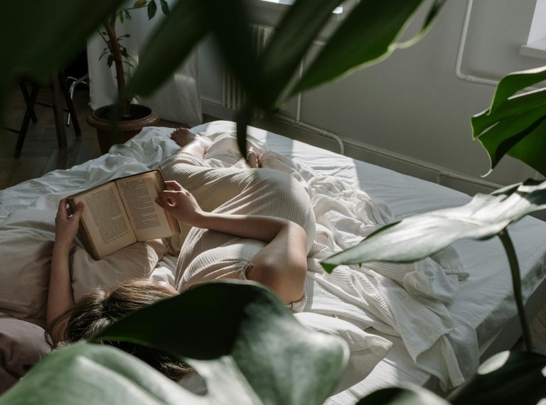 A woman lies in bed reading a book, surrounded by greenery and natural light.