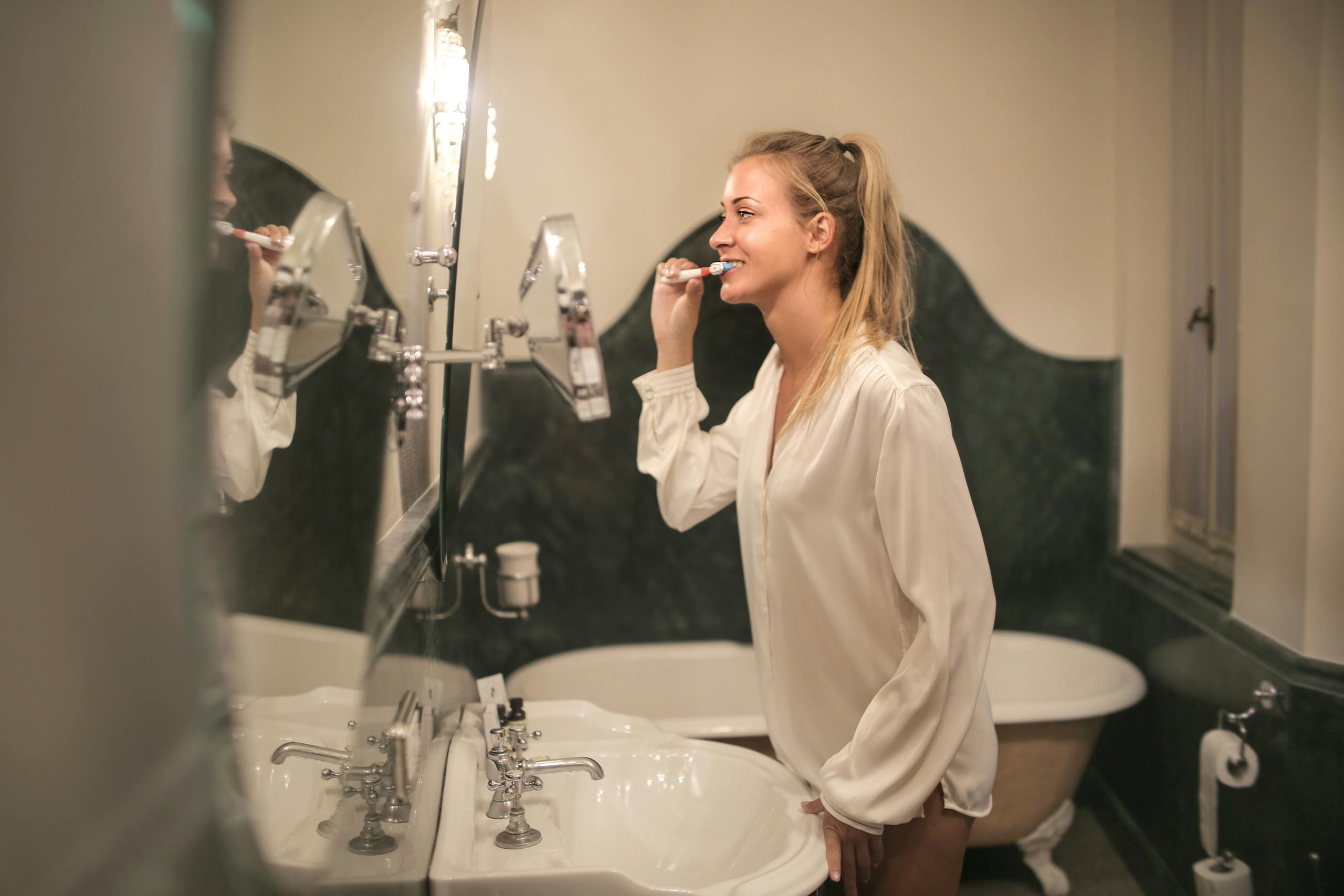 A cheerful young woman in a bathroom brushing her teeth, reflected in a mirror.